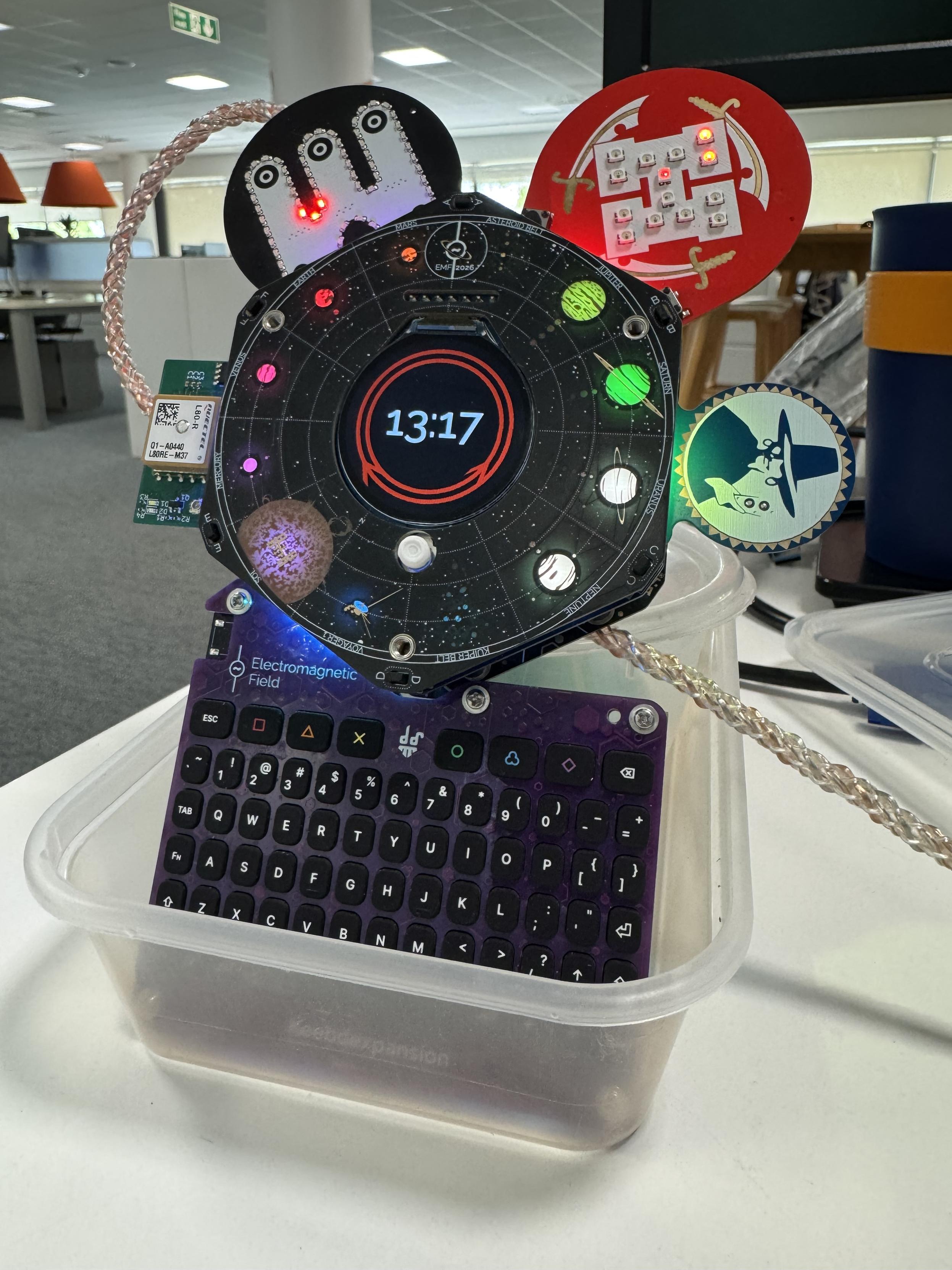 EMF camp spaceagon badge, with national rail clock displayed, and hexpansions including gps, caffeine jitters, essex hackerspace and makerspace, also the keyboard